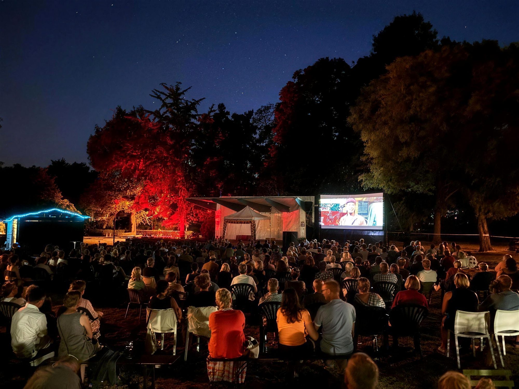 Filmfest im nächtlichen Kronepark: viele Menschen vor der großen Leinwand, die Bäume rot angestrahlt